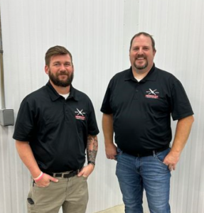 Two men in black polo shirts smiling and standing indoors.