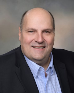 Bald man in a black suit with a blue shirt smiling for a headshot.